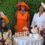 Five women posing for a photo and smiling against a greenery background.