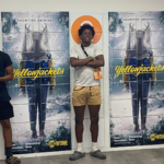 Three boys posing for a photo next to large posters for the tv show &quot;Yellowjackets&quot;.